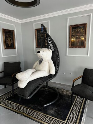 Una stanza con un grande orso di peluche bianco su una sedia a dondolo nera, due sedie nere, un tappeto nero e dorato e tre quadri appesi alle pareti.