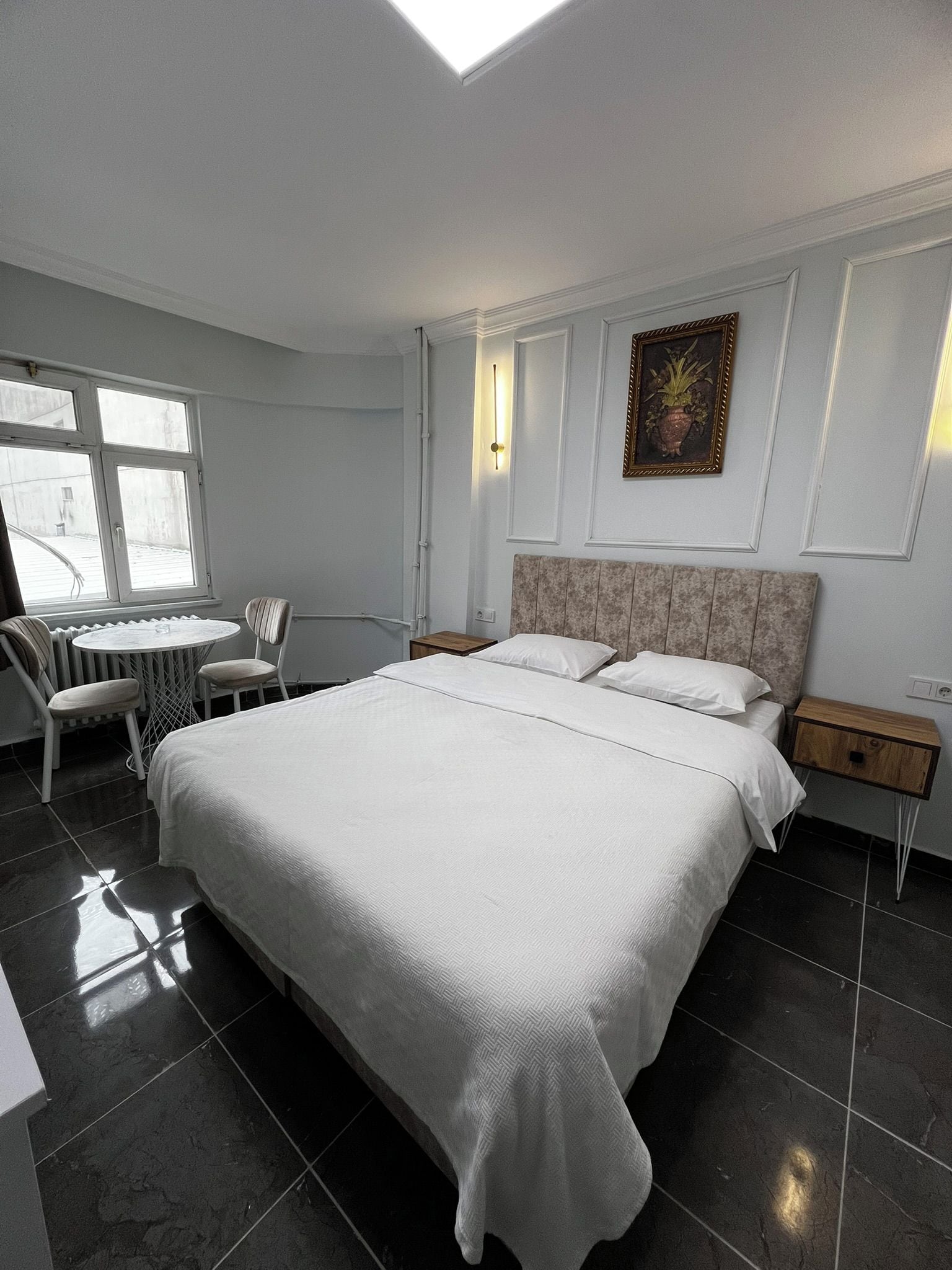 A hotel room with a large bed, a small table, two chairs, a radiator, and a window.