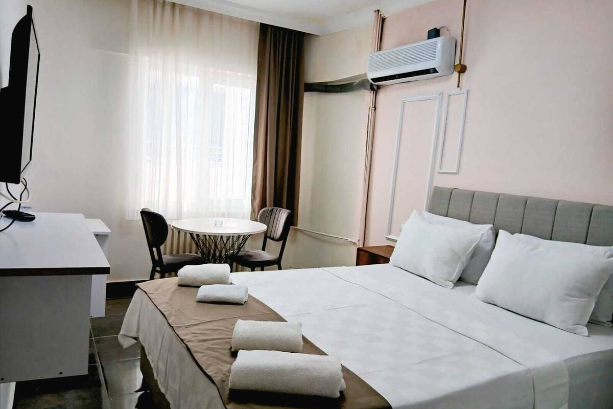 A hotel room with a bed, a desk, a chair, a small table, and an air conditioning unit.