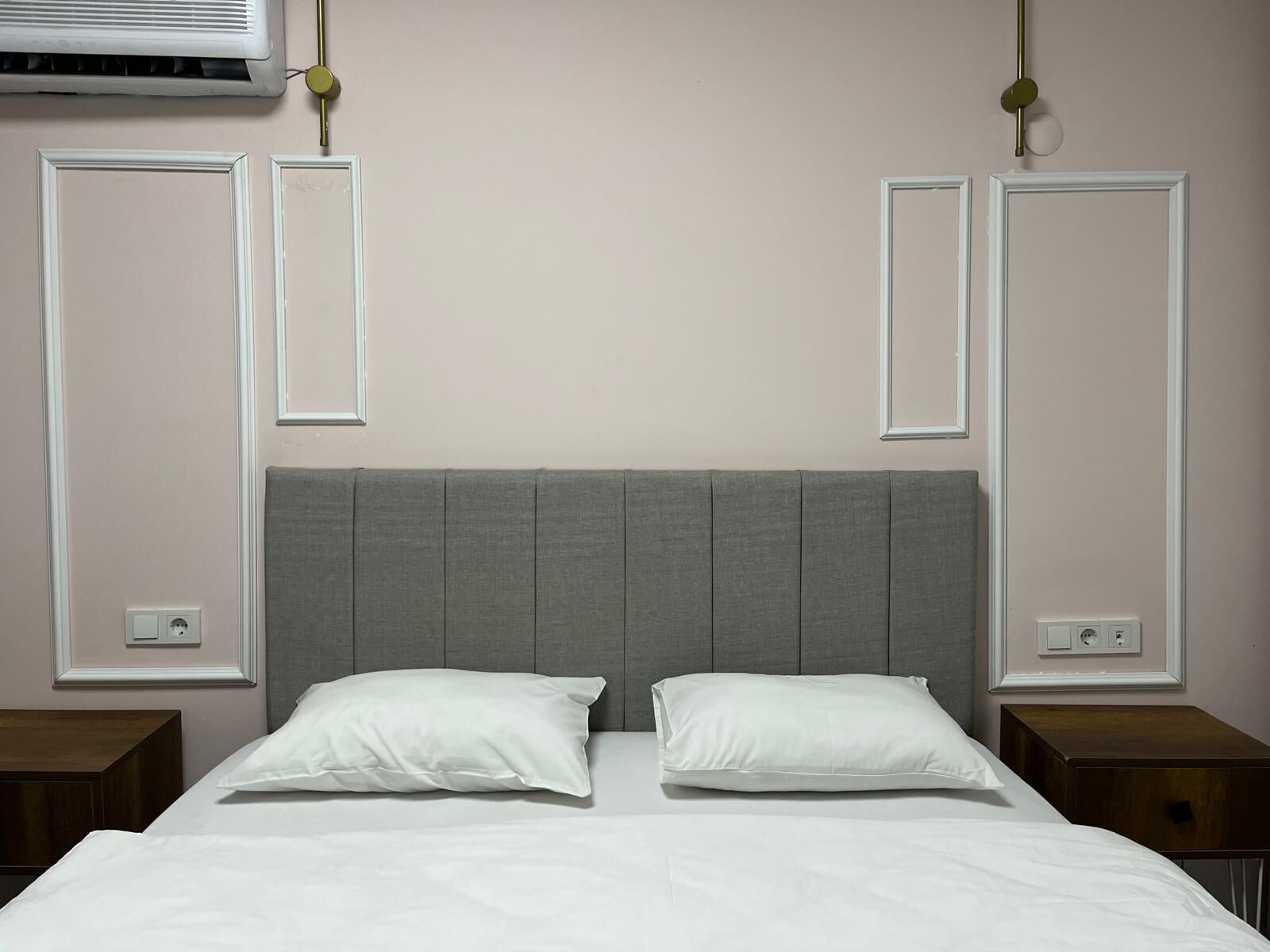 Quarto com cabeceira cinza, roupa de cama branca e duas mesas de cabeceira.
