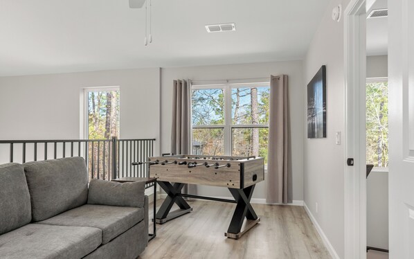 A modern living room with a grey sofa, a wooden foosball table, and large windows offering a view of trees.