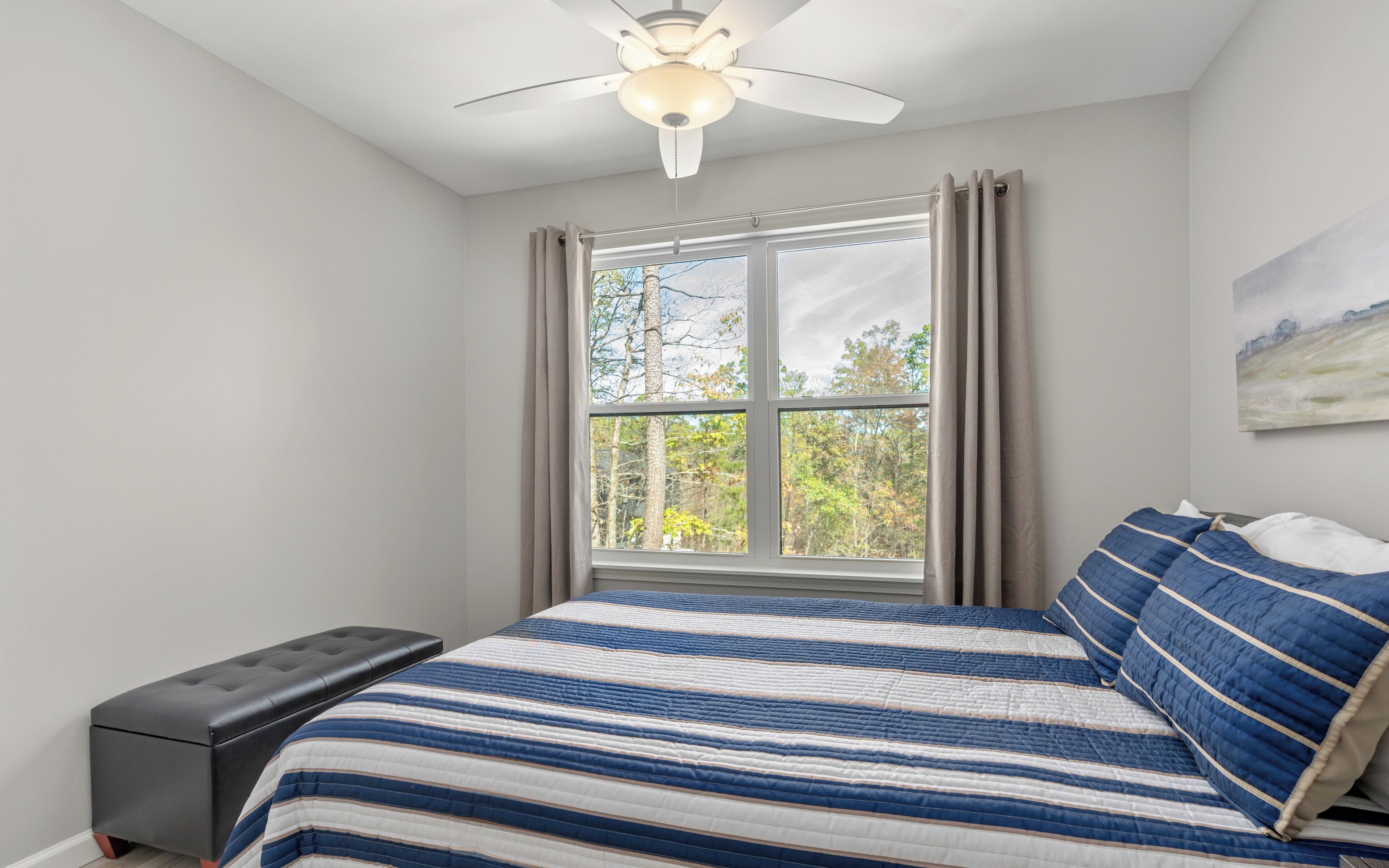 A bedroom with a bed, a bench, a ceiling fan, a window with curtains, and a painting on the wall.