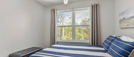 A bedroom with a bed, a bench, a ceiling fan, a window with curtains, and a painting on the wall.