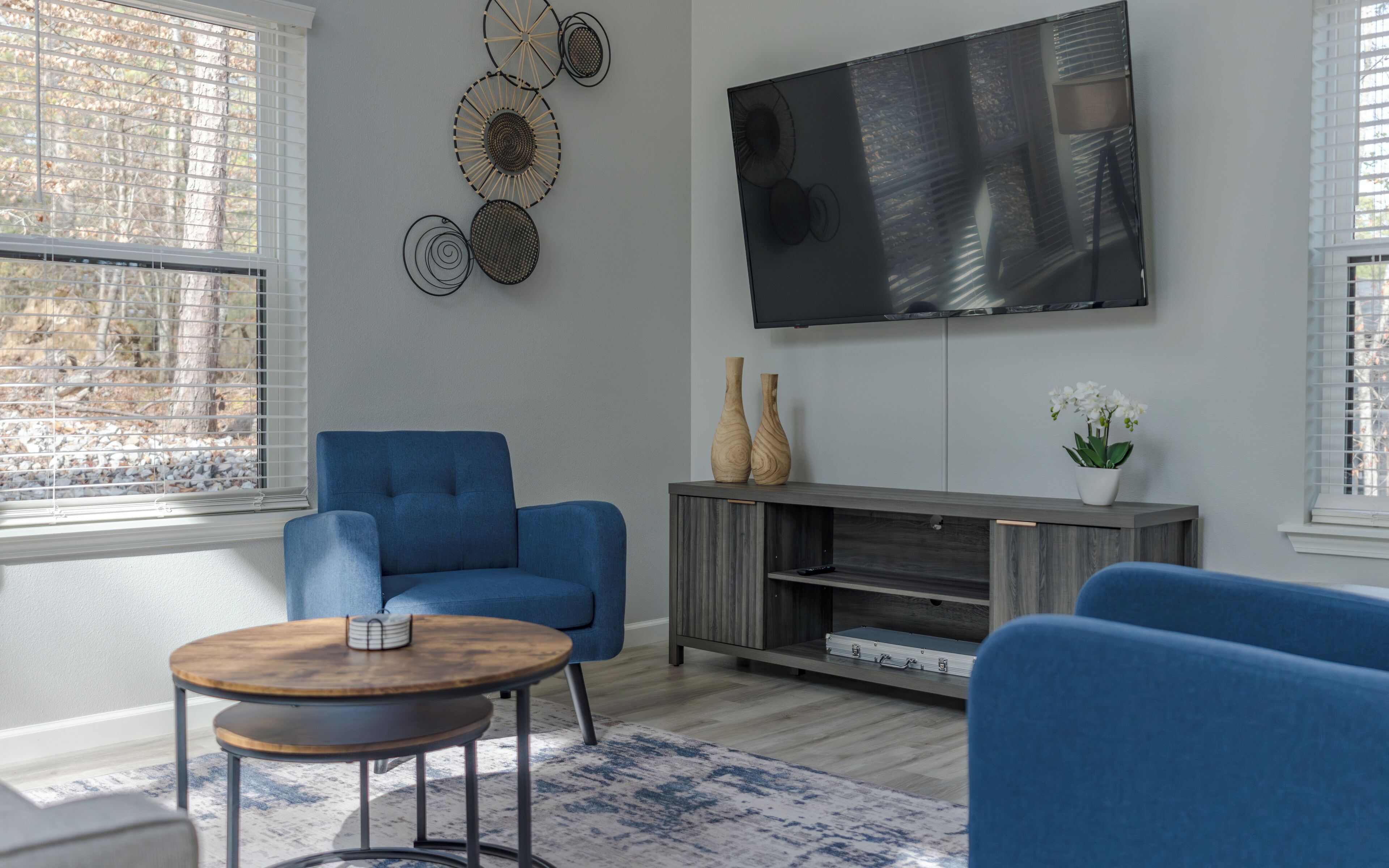 Un salon moderne avec un canapé bleu, une table basse ronde en bois, un meuble de rangement gris et un téléviseur à écran plat fixé au mur.