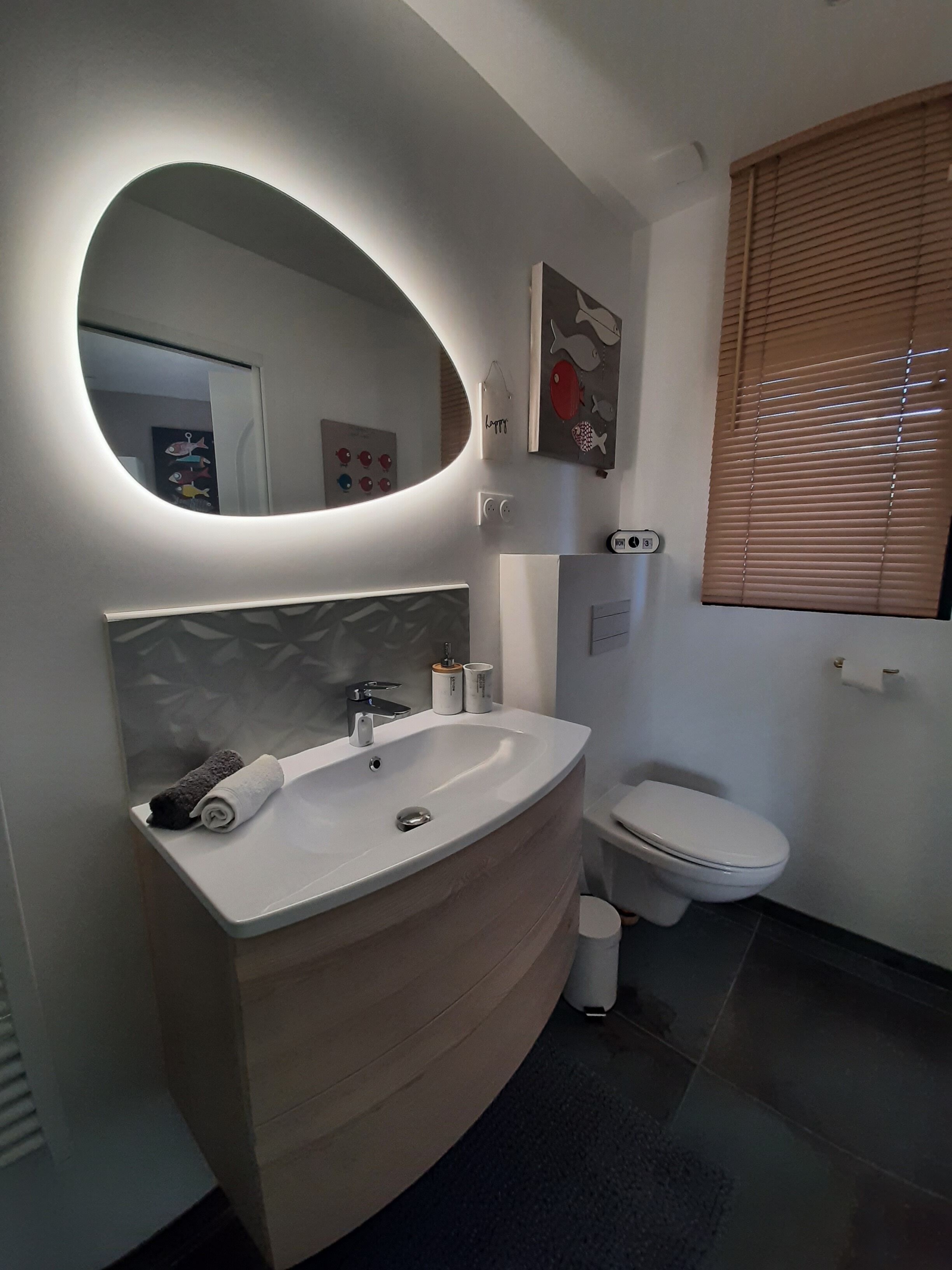 A modern bathroom with a curved vanity, a round mirror, a toilet, and a window with blinds.