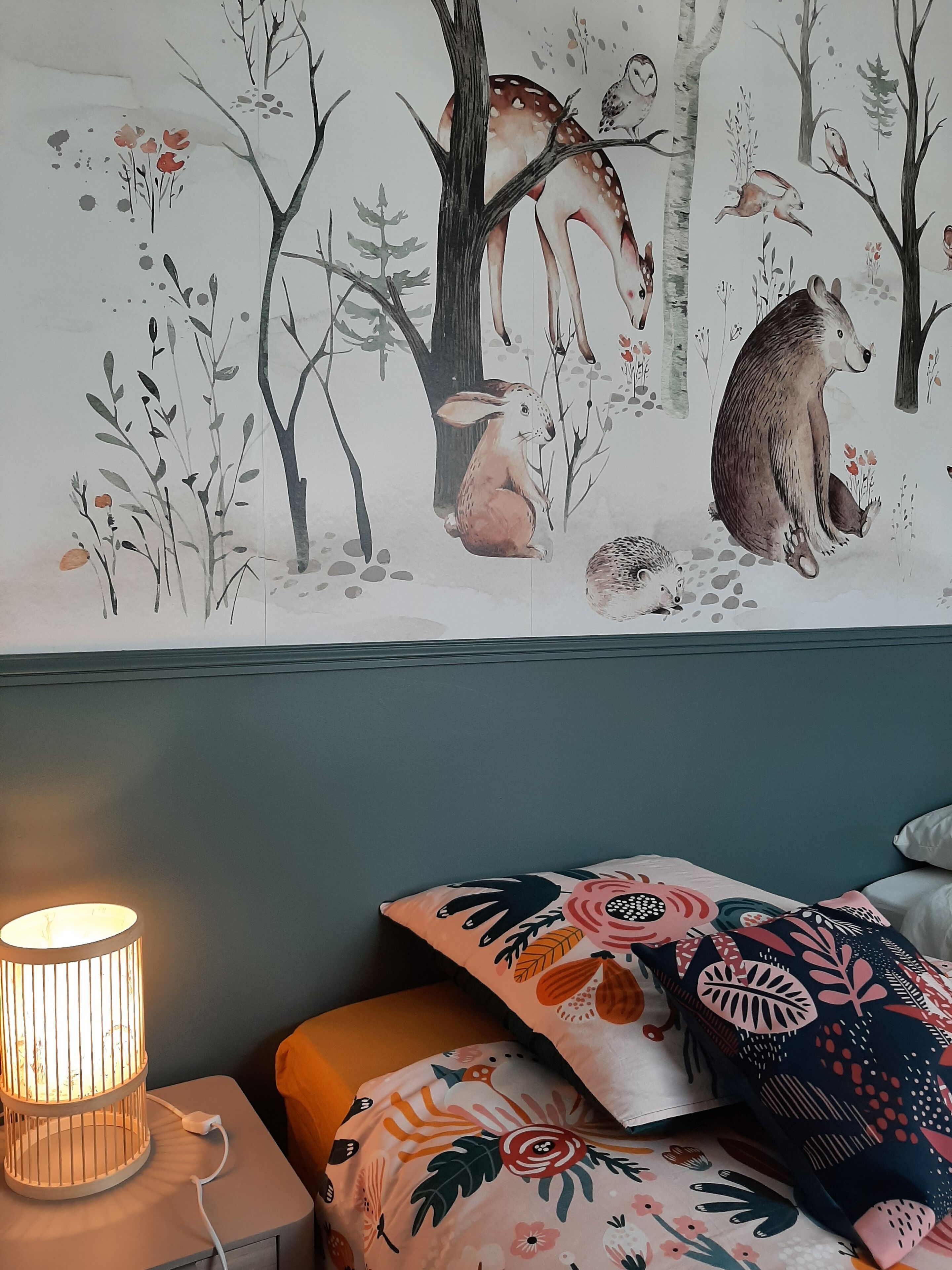 Makuuhuone, jonka seinällä on metsäeläimiä esittävä seinämaalaus, yöpöydällä on lamppu ja sängyllä on kuvioituja tyynyjä.