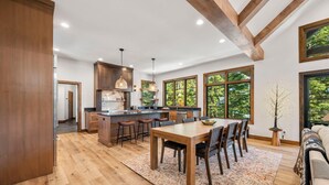 A spacious kitchen with wooden beams, a dining area with a large table and chairs, and large windows offering a view of greenery. - The Tree House | Exclusive Tamarack Perks (Tamarack)