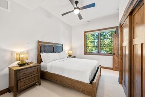 A bedroom with a wooden bed, two bedside tables with lamps, a ceiling fan, and a window with a view of trees. - The Tree House | Exclusive Tamarack Perks (Tamarack)