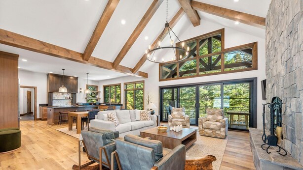 A spacious living area with a large sofa, leather chairs, a coffee table, and a stone fireplace. There is a kitchen in the background with wooden beams and large windows. - The Tree House | Exclusive Tamarack Perks (Tamarack)