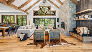 A spacious living area with a high ceiling, stone fireplace, and large windows offering views of greenery. - The Tree House | Exclusive Tamarack Perks (Tamarack)
