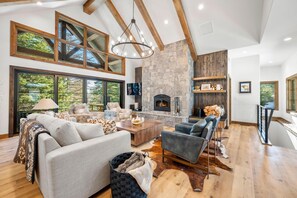 A spacious living area with a stone fireplace, large windows, and a high ceiling with exposed beams.