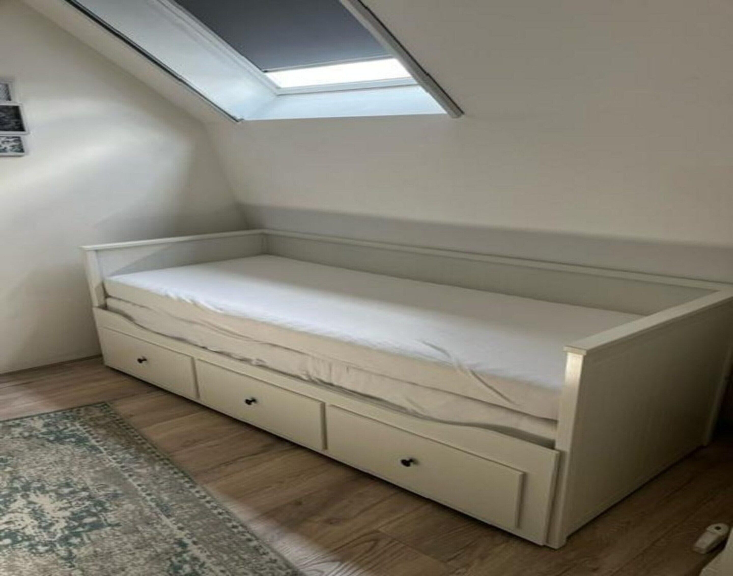 Ein kleines Zimmer mit einem einzelnen Bett, einem Oberlicht, einem gemusterten Teppich und Holzboden.