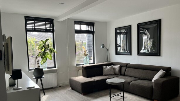 A modern living room with a grey sectional sofa, a round coffee table, and two windows with black blinds. - Holiday Home Near Dunes, Beach, sea (Sint Maartensvlotbrug)