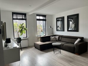 A modern living room with a grey sectional sofa, a round coffee table, and two windows with black blinds. - Holiday Home Near Dunes, Beach, sea (Sint Maartensvlotbrug)