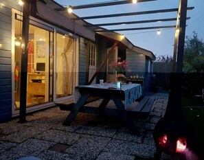 An outdoor patio area with a table and chairs, a wooden deck, and string lights. - Attractive Bungalow Near Beach, Dunes (Sint Maartensvlotbrug)