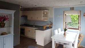 A kitchen with wooden cabinets, a white countertop, and a dining area with a table and chairs. - Attractive Bungalow Near Beach, Dunes (Sint Maartensvlotbrug)