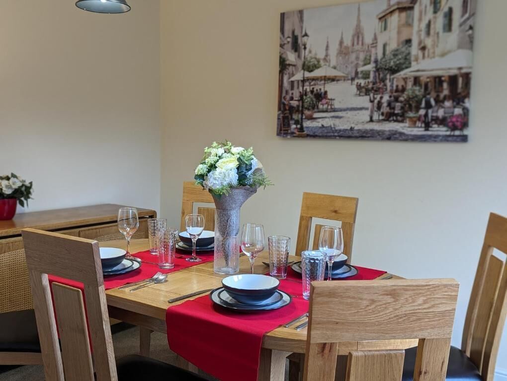 Une table à manger dressée avec des assiettes, des verres et des couverts, un vase rempli de fleurs et une nappe rouge.