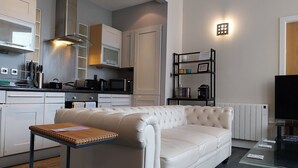 A modern kitchen with white cabinets, a microwave, and a coffee maker. There is a white leather sofa with a small table in front. A TV is on the right side of the room.