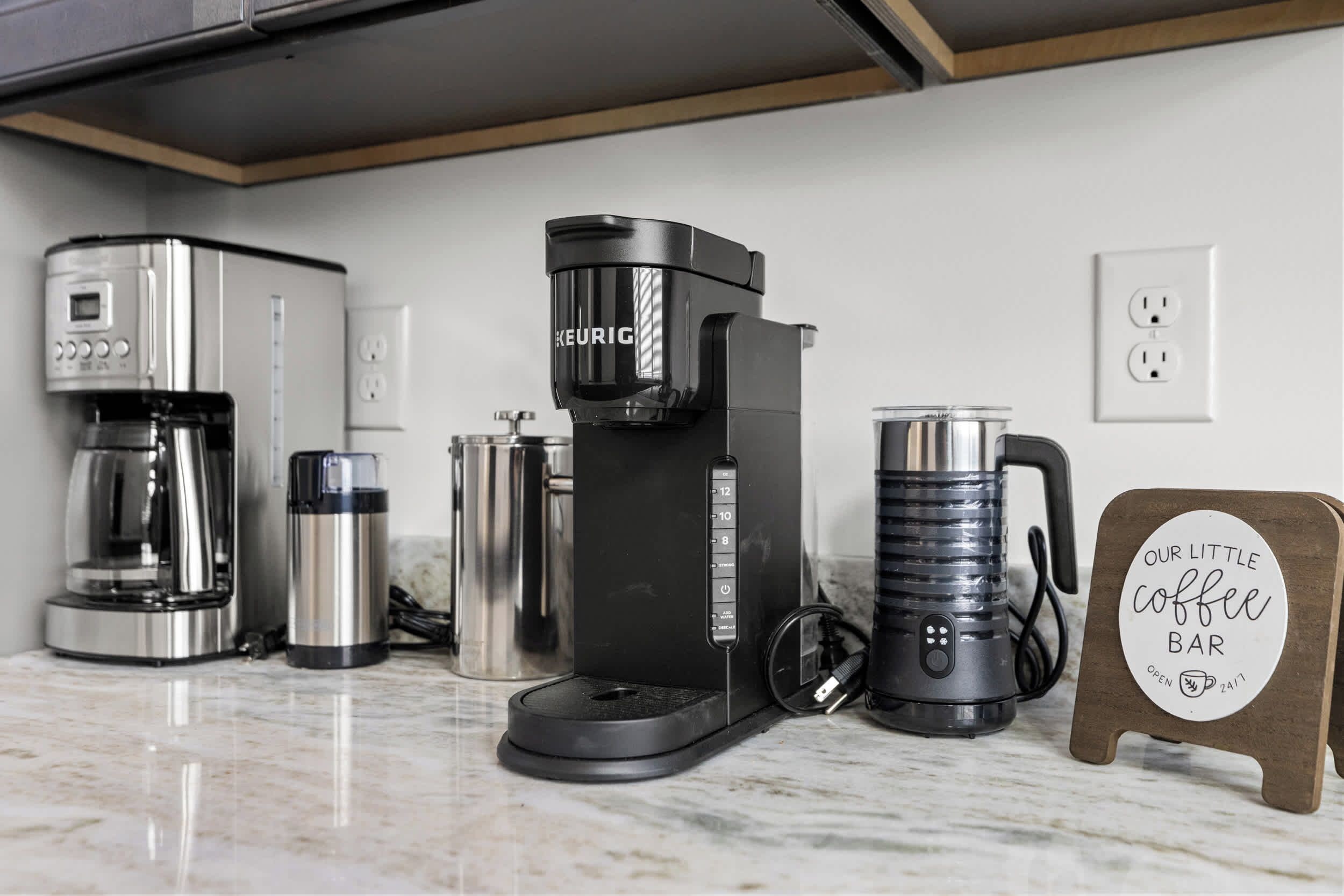 Uma cozinha moderna com cafeteira Keurig, jarra de café e um letreiro que diz 'Nosso Pequeno Café'.
