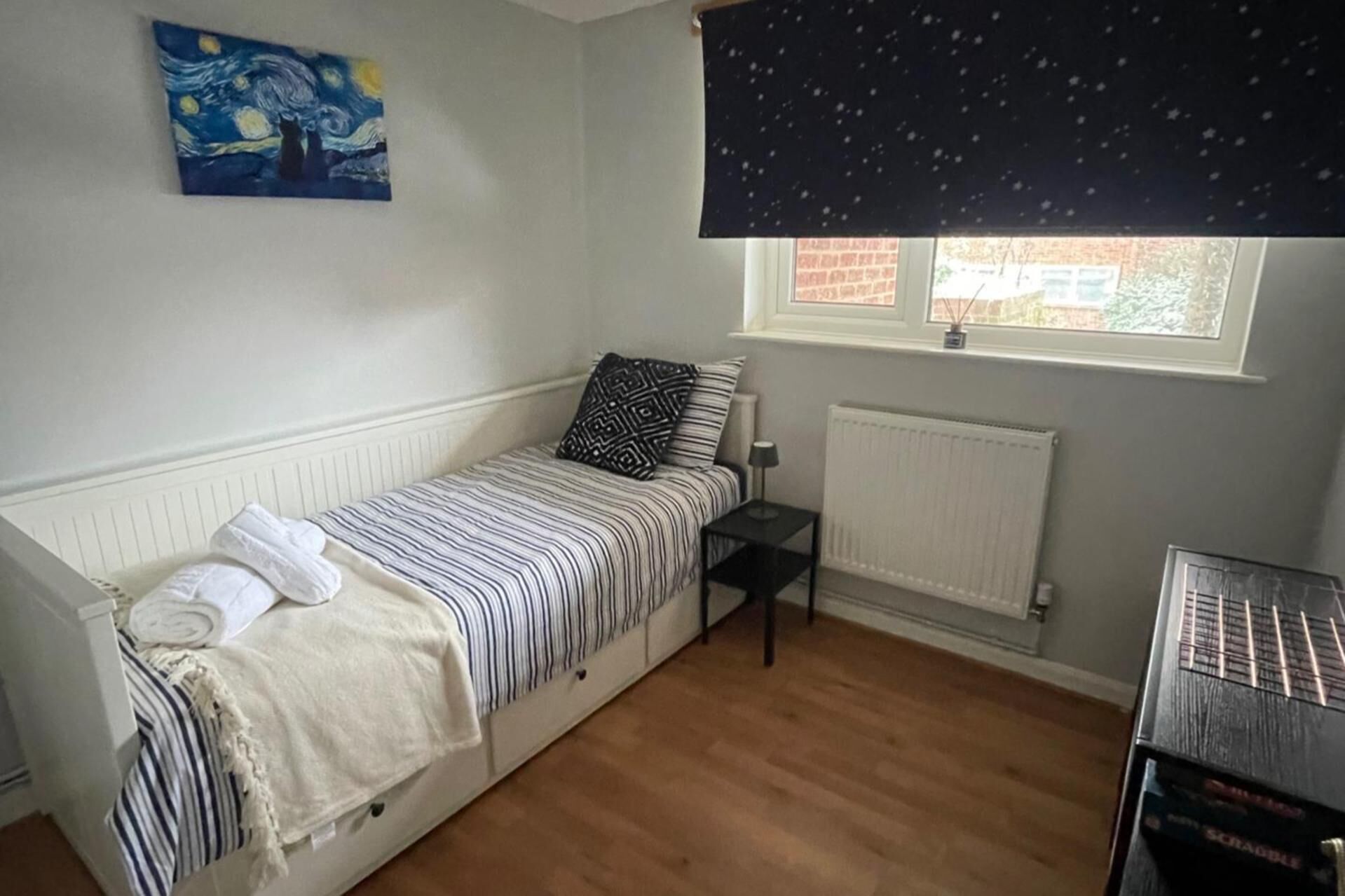 A single bed with a striped bedspread, a pillow, a bedside table with a lamp, a radiator, a window with a curtain, and a painting on the wall.
