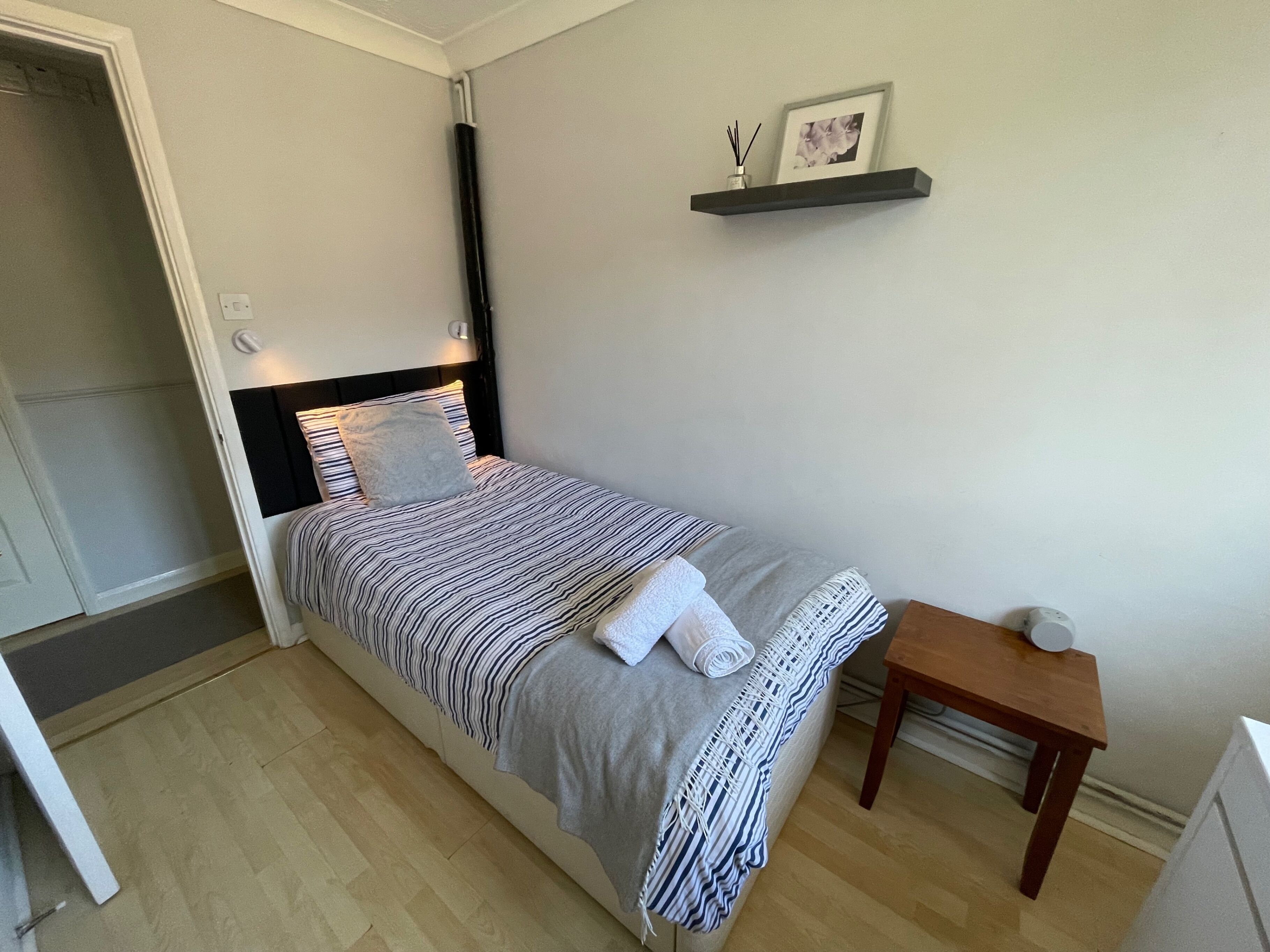 Ein kleines, ordentlich eingerichtetes Schlafzimmer mit Bett, Nachttisch und einem Regal mit einem Bild und einer Vase.