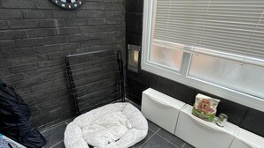 Une pièce d’angle avec un mur en briques noires, un radiateur blanc, un panier pour animaux de compagnie et une fenêtre avec des stores.