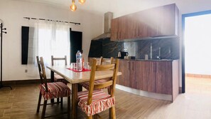 A dining area with a wooden table and chairs, a kitchen with dark cabinets, and a window with curtains. - Apartment Alegria in the Stunning Village of Mozaga, Lanzarote (Teguise)