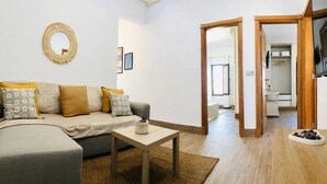 Una sala de estar con sofá, mesa de centro y televisor. Hay una puerta que da a un baño y una zona de cocina.