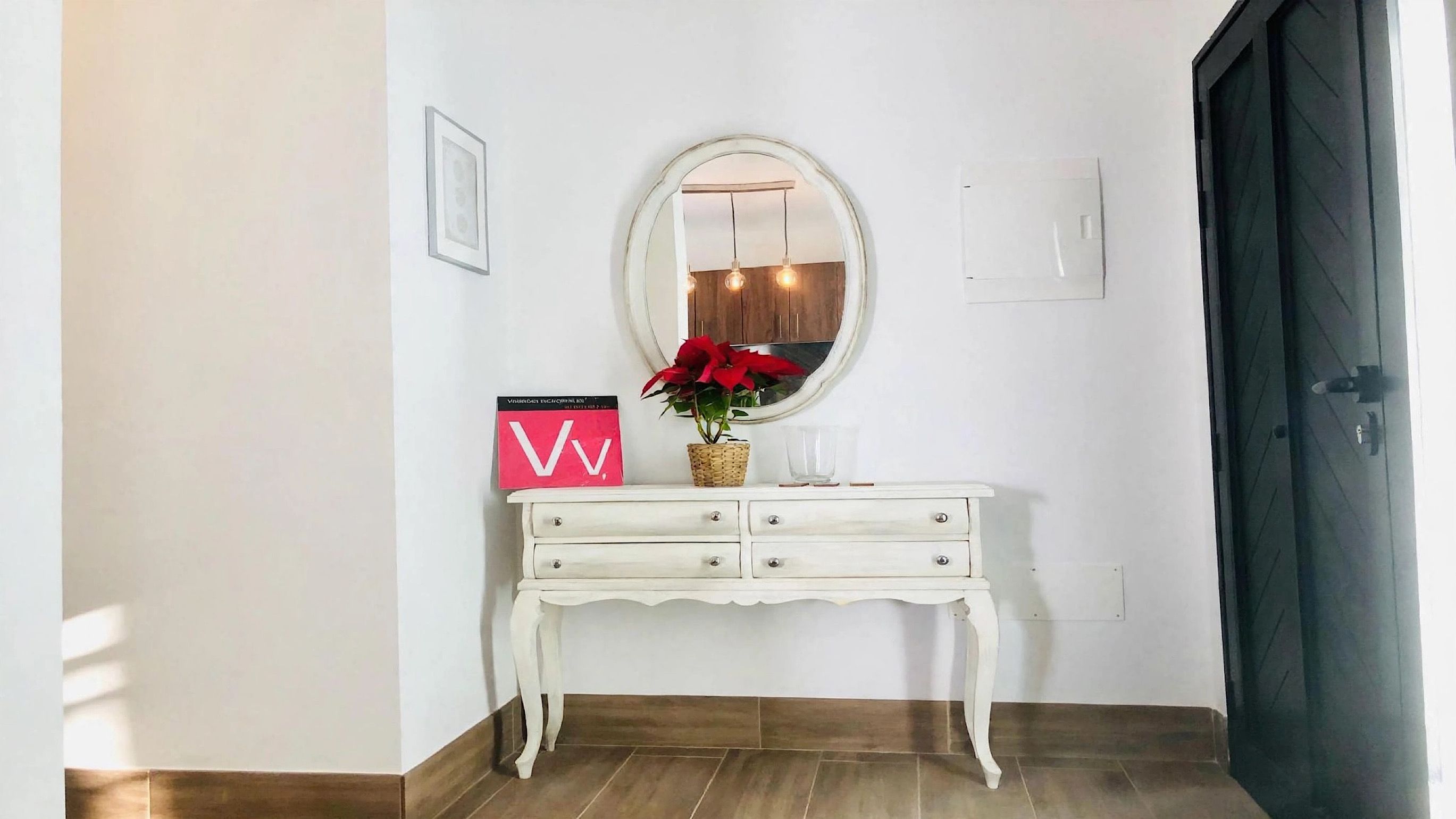 A white console table with drawers, a mirror, a red sign with a white 'V', and a potted plant.