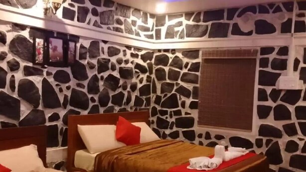 Habitación de hotel con diseño de pared de piedra, dos camas con ropa de cama roja y blanca, un ventilador de techo y suelo a cuadros.