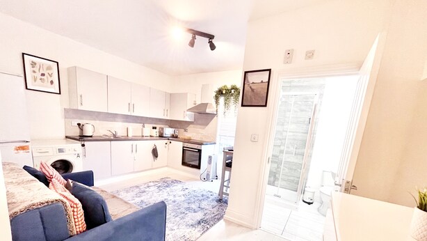 A modern kitchen with white cabinets, a built-in oven, and a microwave. There is a blue sofa with a patterned throw blanket in the living area. - 25min to Central - Zone 2 - Free Parking - Sleep 6 (London)