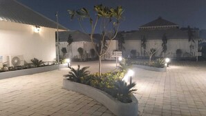 Un patio con suelo de baldosas, una jardinera central con flores amarillas y un edificio con techo abovedado al fondo.