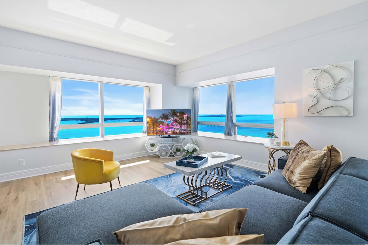 A modern living room with a large TV, a yellow chair, a sofa, and a view of the ocean through large windows.