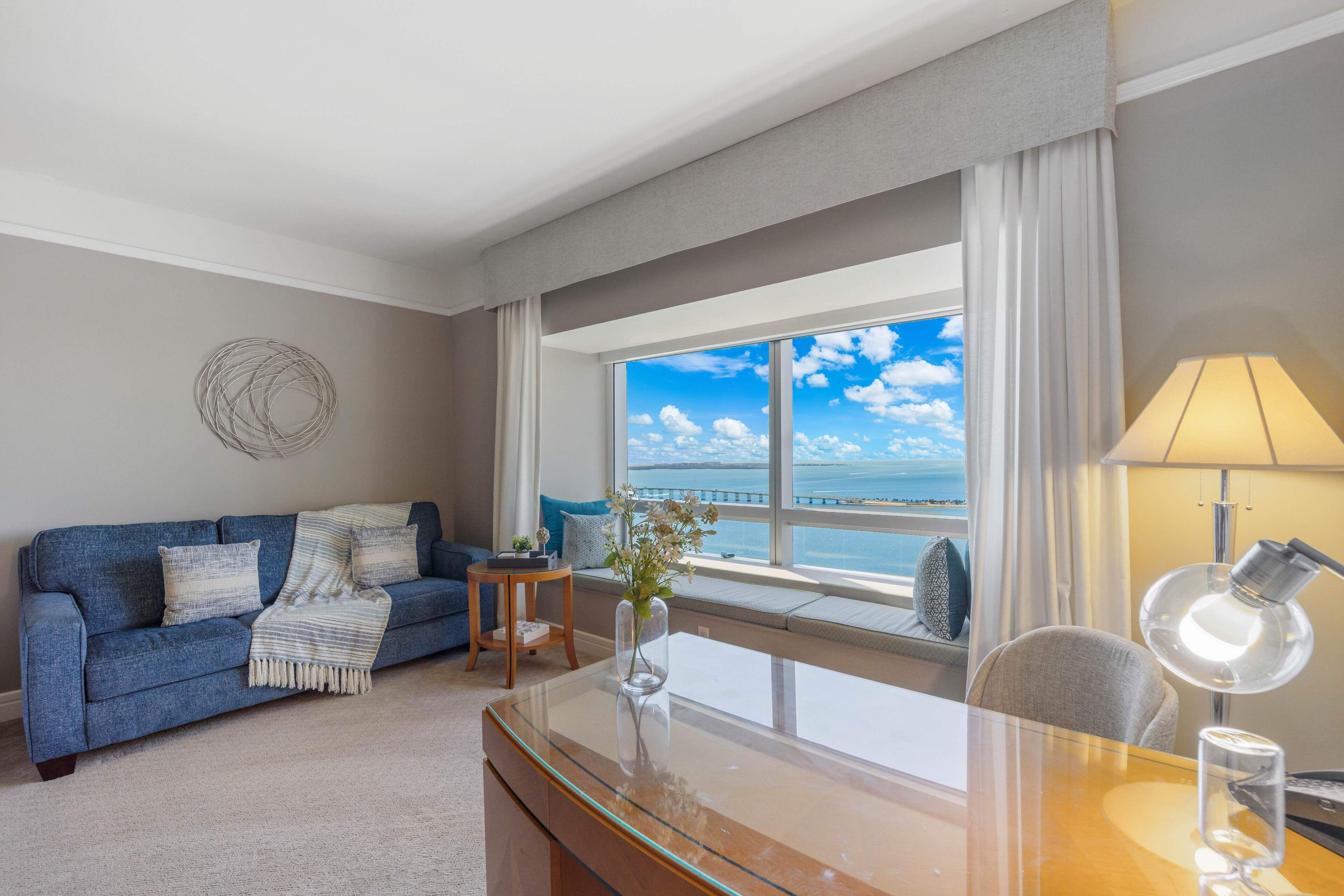 Een moderne hotelkamer met een groot raam dat uitzicht biedt op zee en een brug, een blauwe bank met decoratieve kussens, een klein bijzettafeltje met een vaas, en een bureau met een lamp.