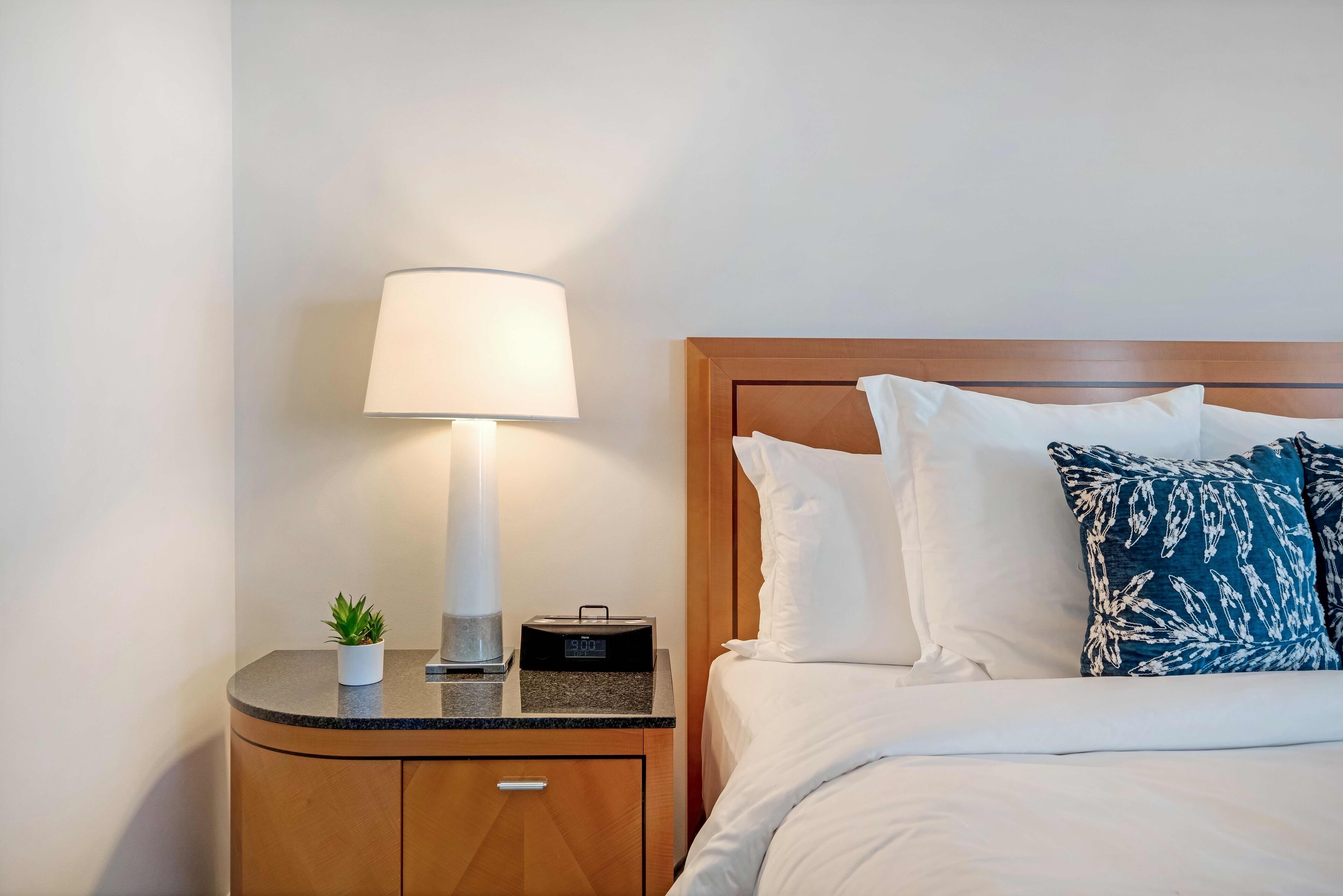 Een net opgemaakt bed met witte en blauwe kussens, een bedlampje, een kleine potplant en een radio op een houten nachtkastje.