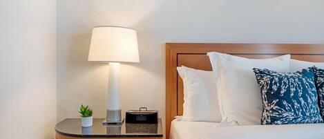 Een net opgemaakt bed met witte en blauwe kussens, een bedlampje, een kleine potplant en een radio op een houten nachtkastje.