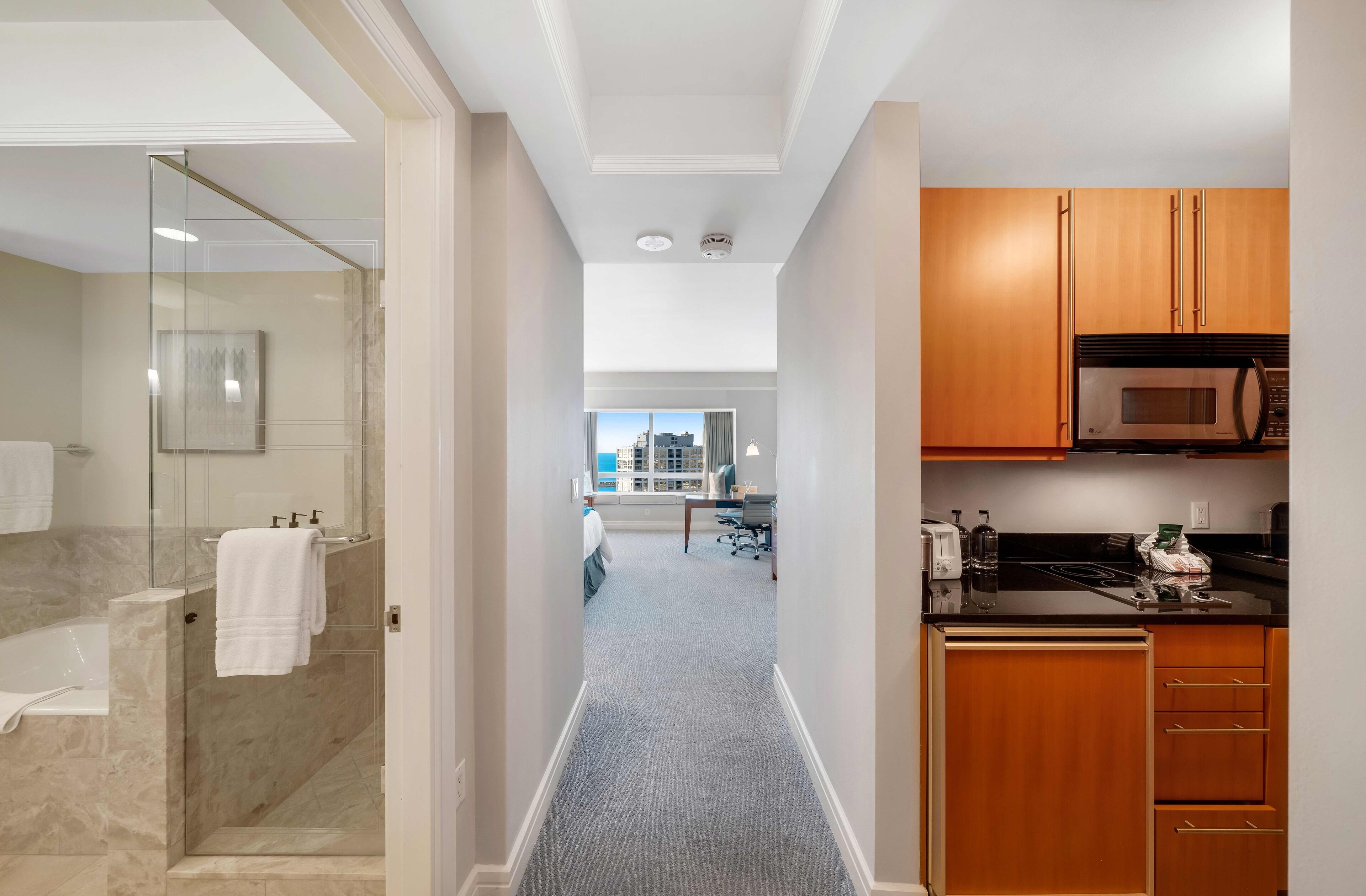 A modern hotel room with a bathroom, a hallway leading to a kitchen, and a view of the city.