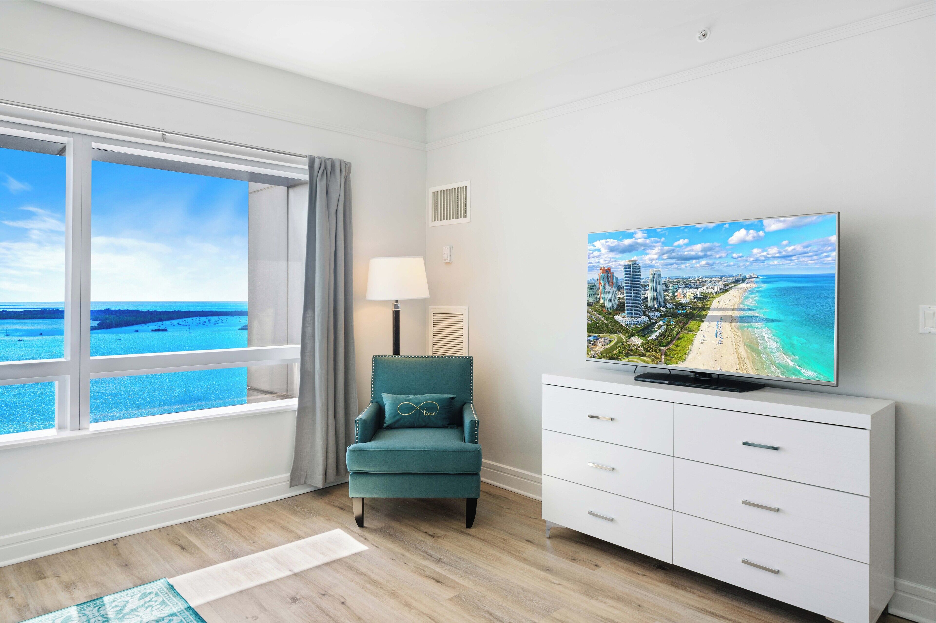 A room with a large window offering a view of the beach and ocean, a teal armchair with a cushion, a white TV stand with a flat-screen TV, and a floor lamp.