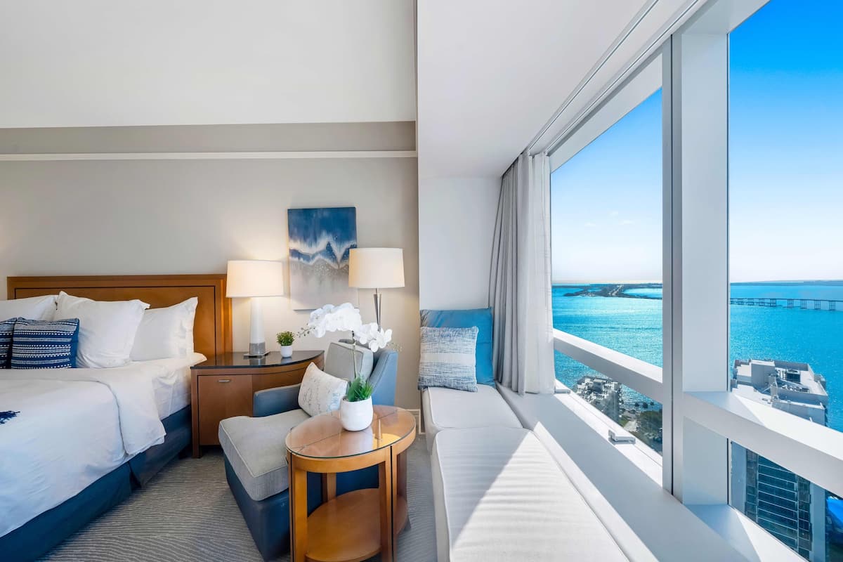 A hotel room with a bed, bedside table, lamp, and a view of the ocean.