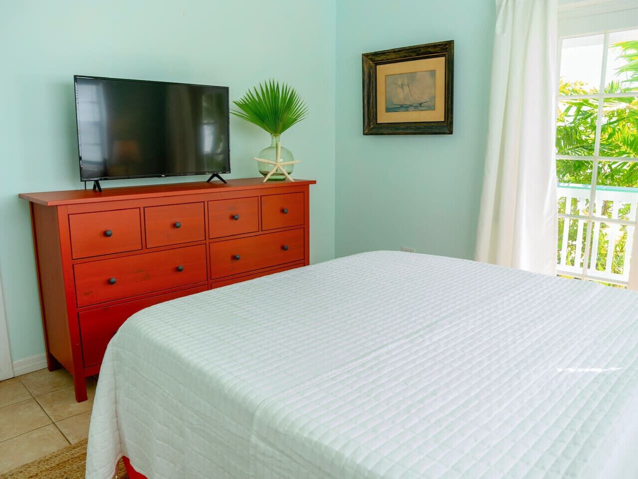 A room with a bed, a red dresser with drawers, a television on top, a framed picture on the wall, and a window with curtains.