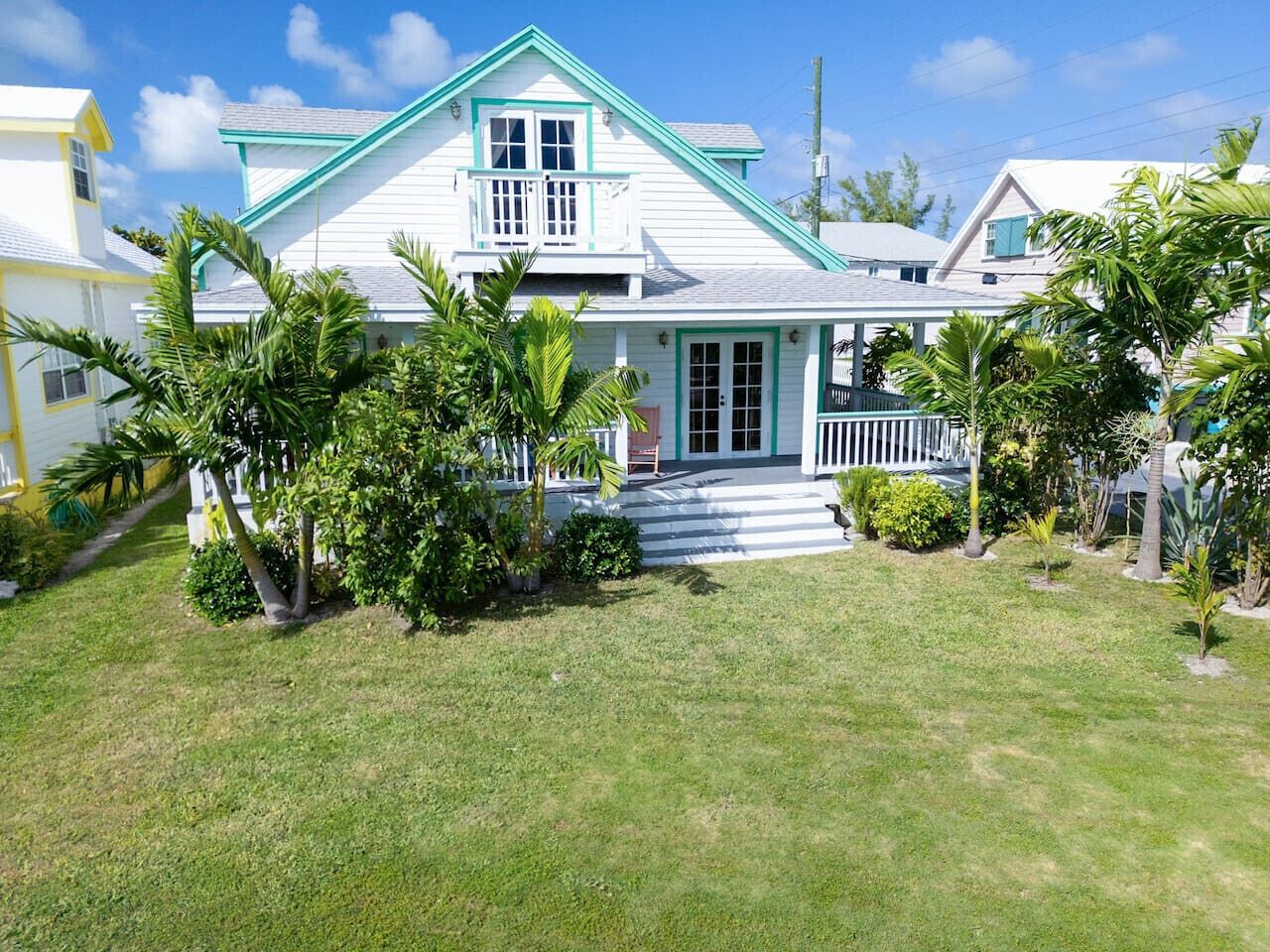 A two-story house with a white exterior and teal trim, surrounded by a well-maintained lawn and various tropical plants.