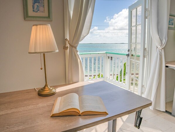A room with a wooden table, an open book, a lamp, and a view of the sea through a balcony.
