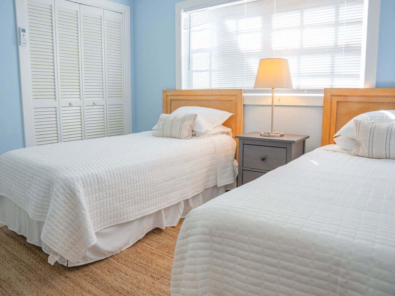 A bedroom with two beds, a bedside table, a lamp, and a window with blinds.