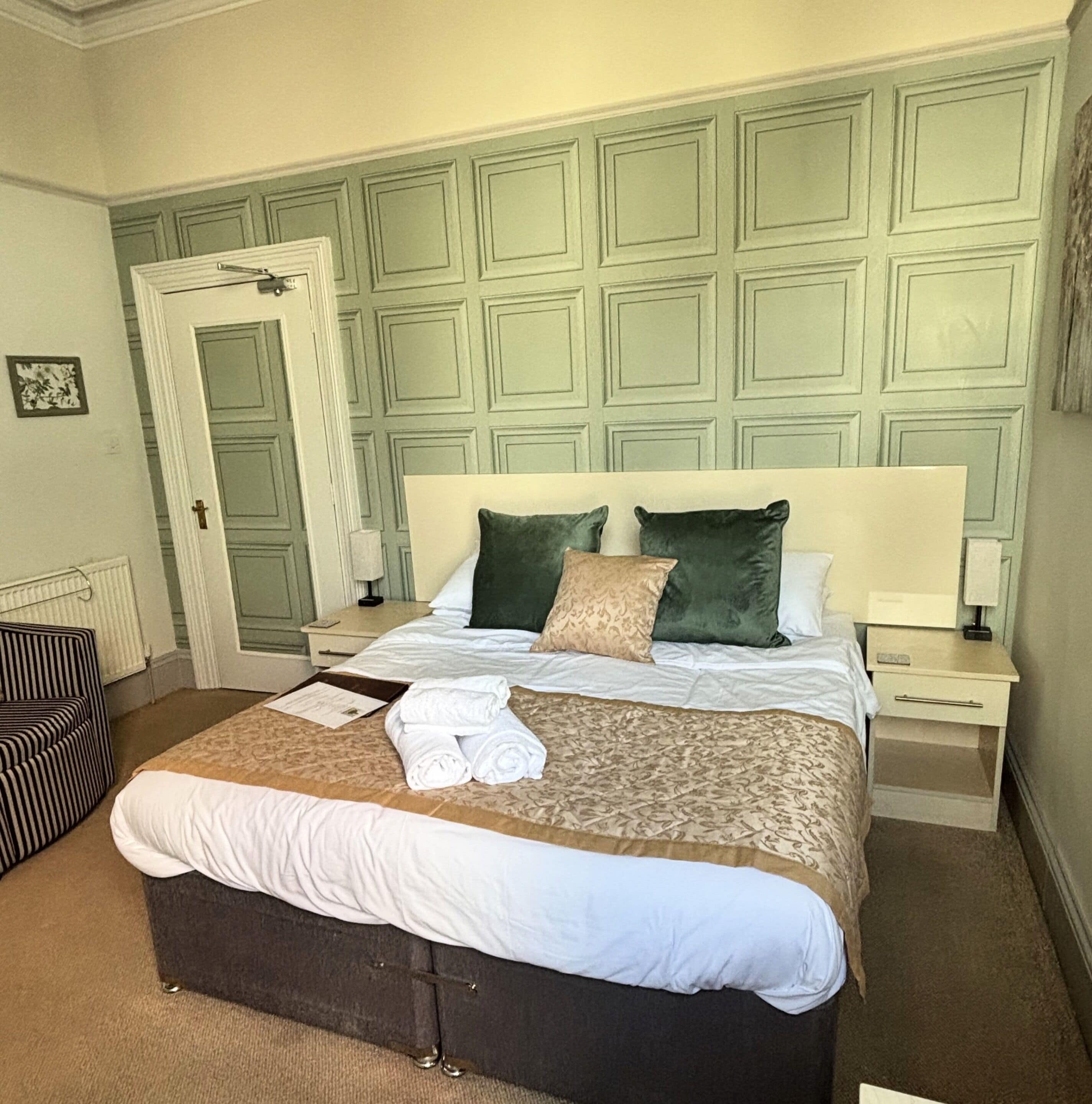 A neatly made bed with a dark headboard, white bedding, and two pillows. There is a bedside table with a lamp and a small drawer. The room has a green paneled wall and a door with a white frame.