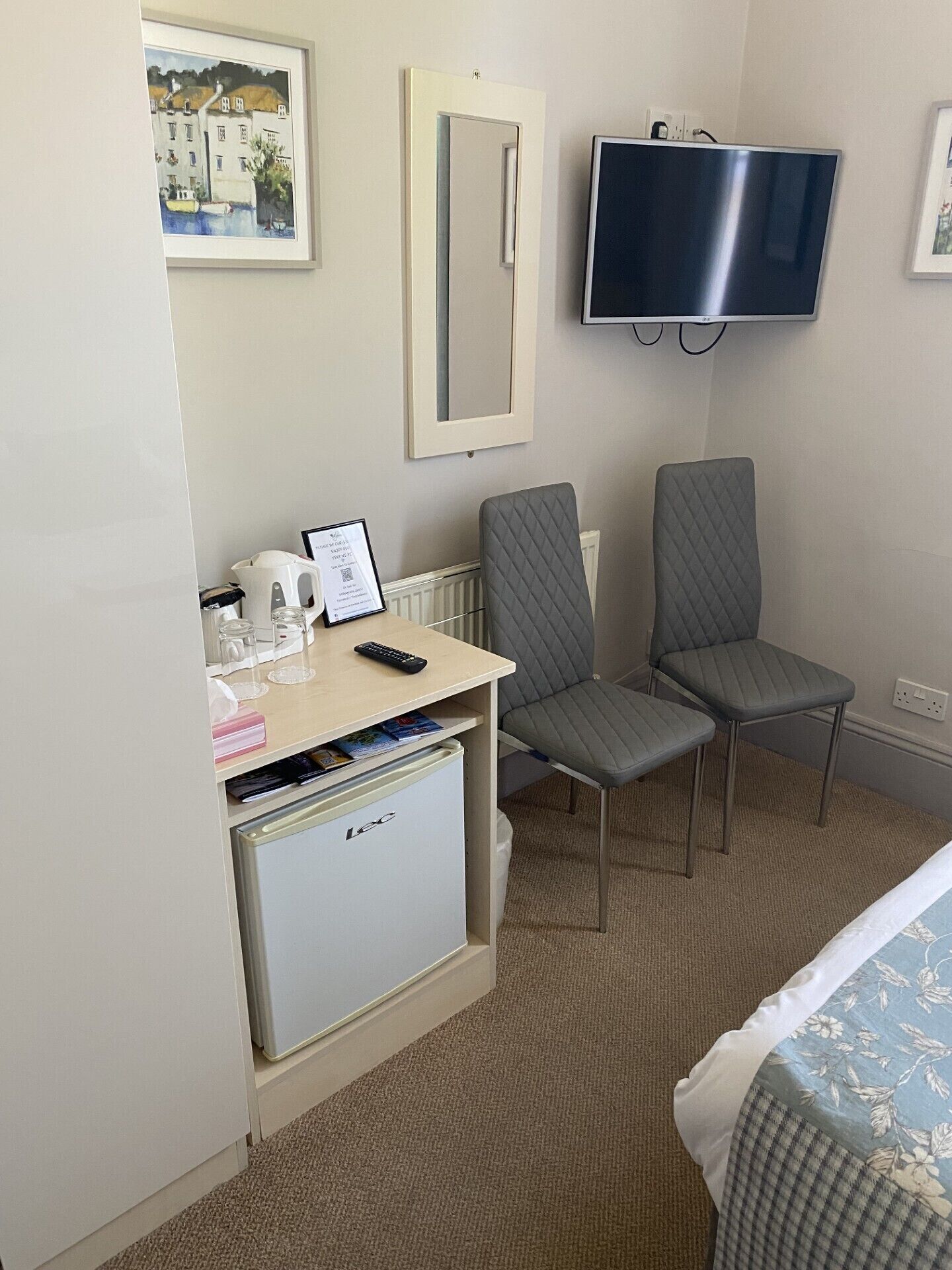 Habitación de hotel con cama, escritorio con refrigerador, dos sillas, televisor y un cuadro en la pared.