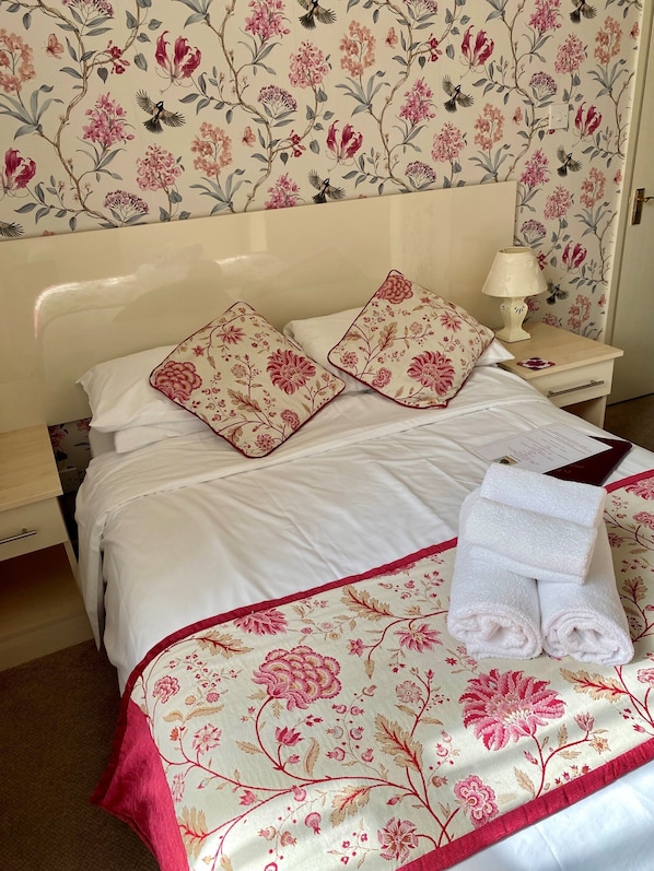 A neatly made bed with floral-patterned pillows and a matching bedspread, a bedside lamp, and a folded towel on the nightstand. - 74 Belgravia (Torquay)