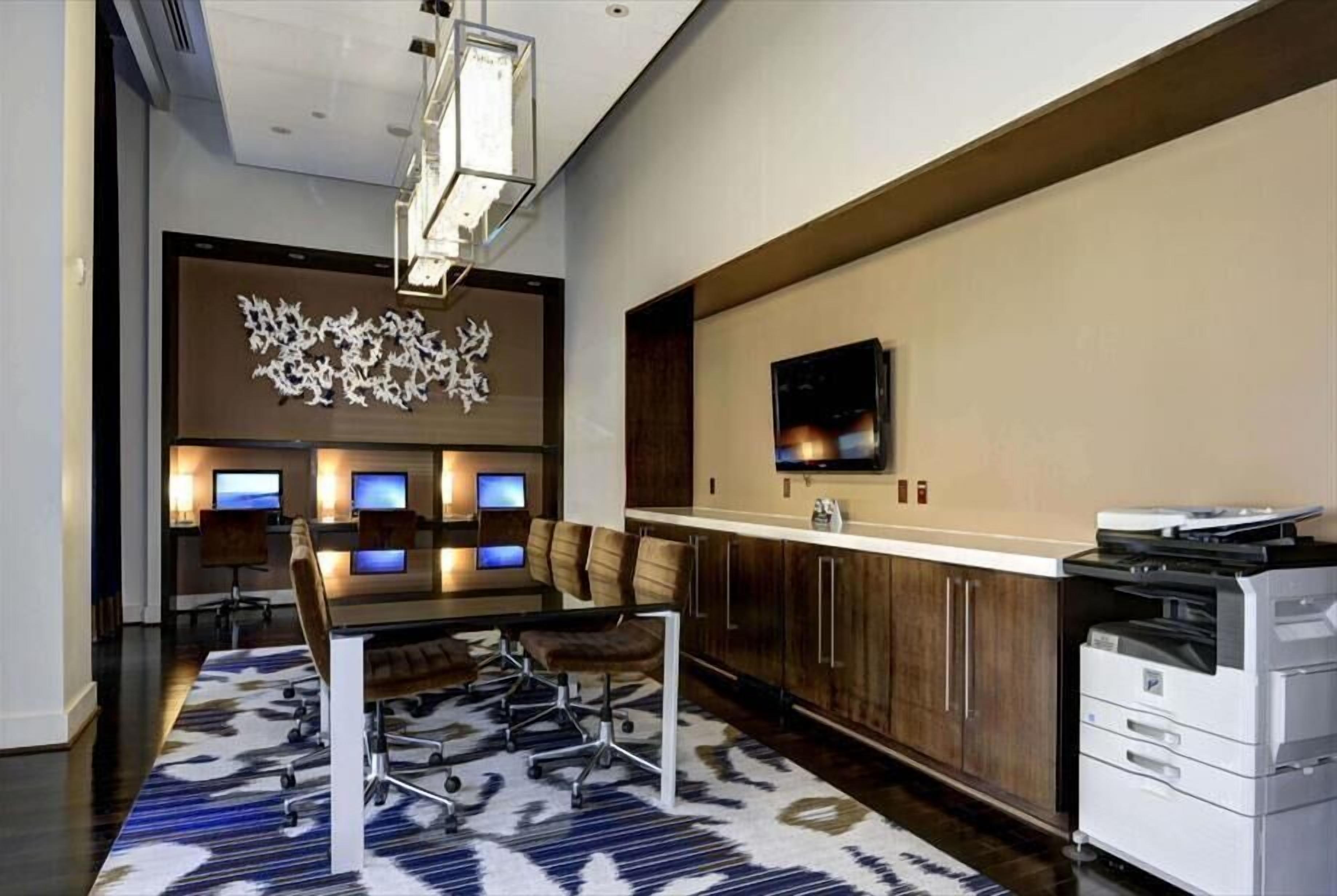 A modern conference room with a long table, multiple chairs, a large abstract art piece, a flat-screen TV, and a printer.