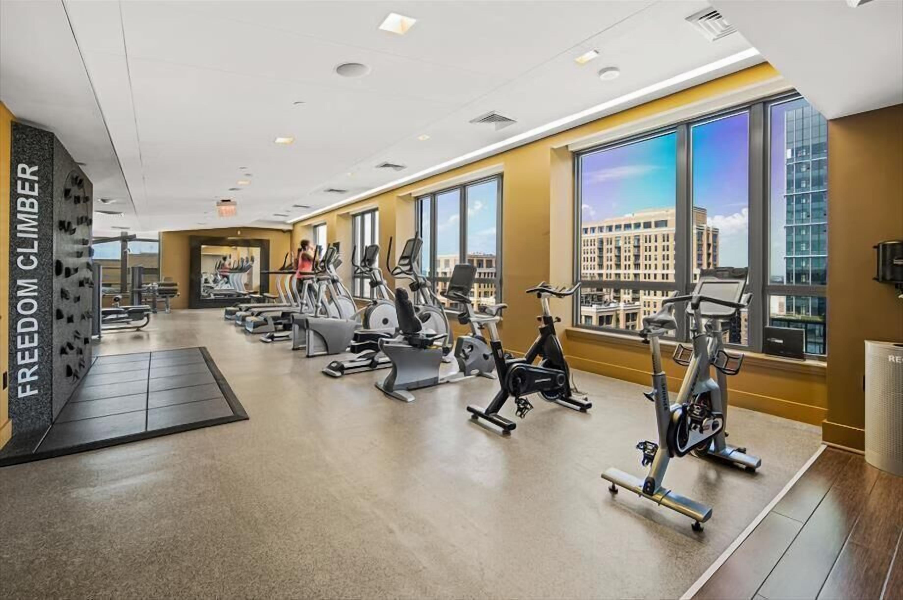 A modern gym with various exercise machines, large windows offering city views, and a prominent "FREEDOM CLIMBER" sign.