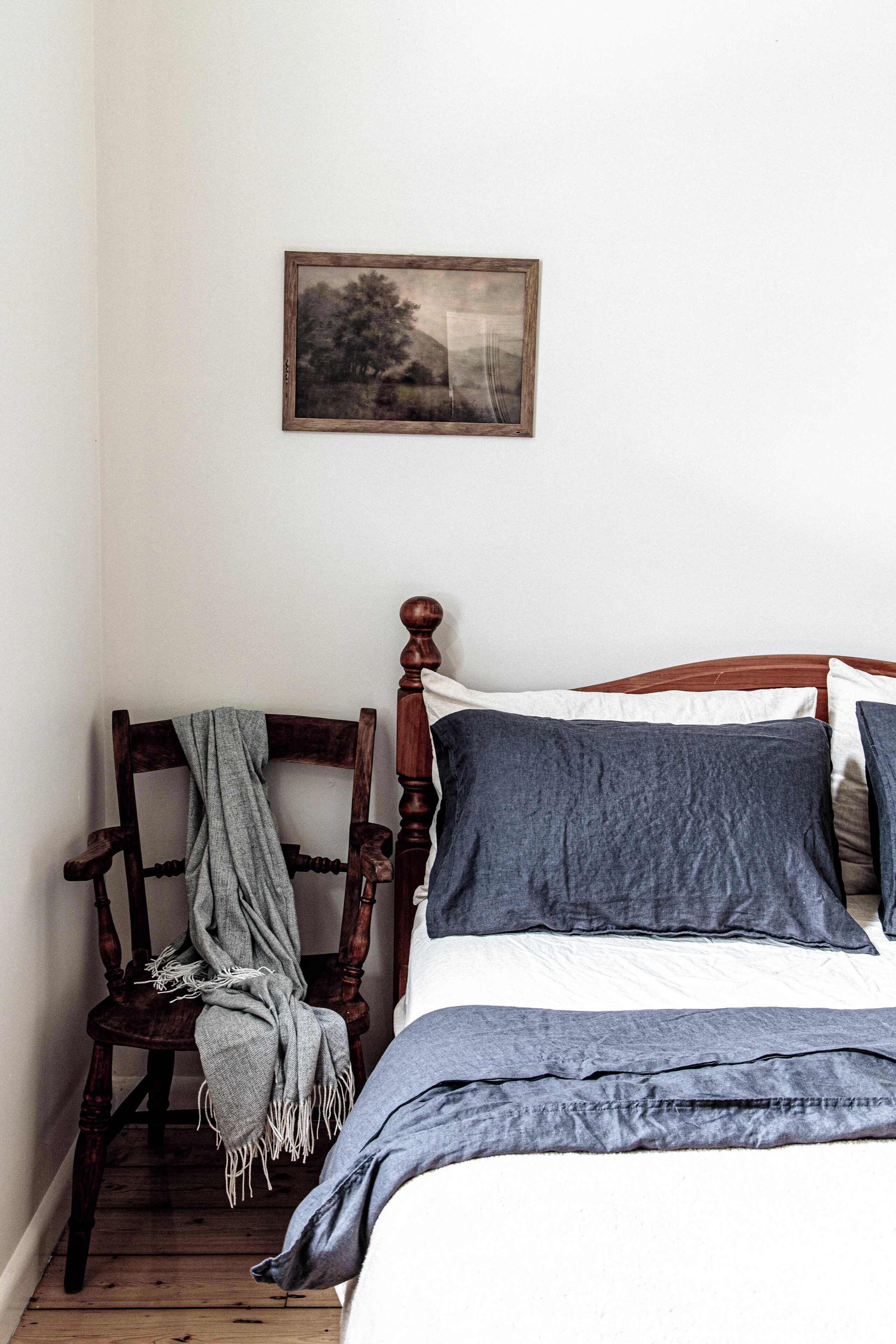 A bedroom with a wooden bed, a chair with a blanket, and a framed picture on the wall.