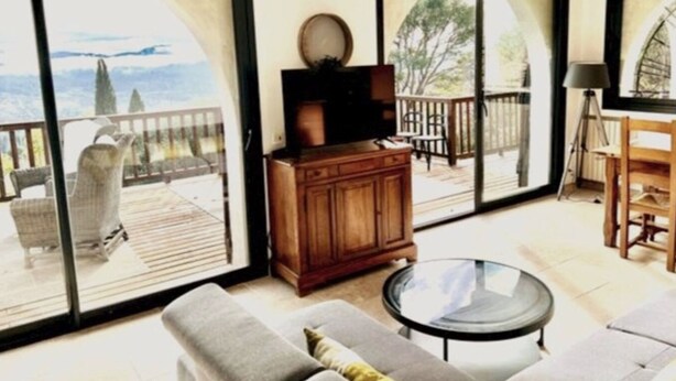 A spacious living area with a sofa, a coffee table, and a television on a wooden cabinet. Large sliding glass doors lead to an outdoor balcony with a view of the mountains. - Le Belvedere-Charme de la Provence (Montauroux)